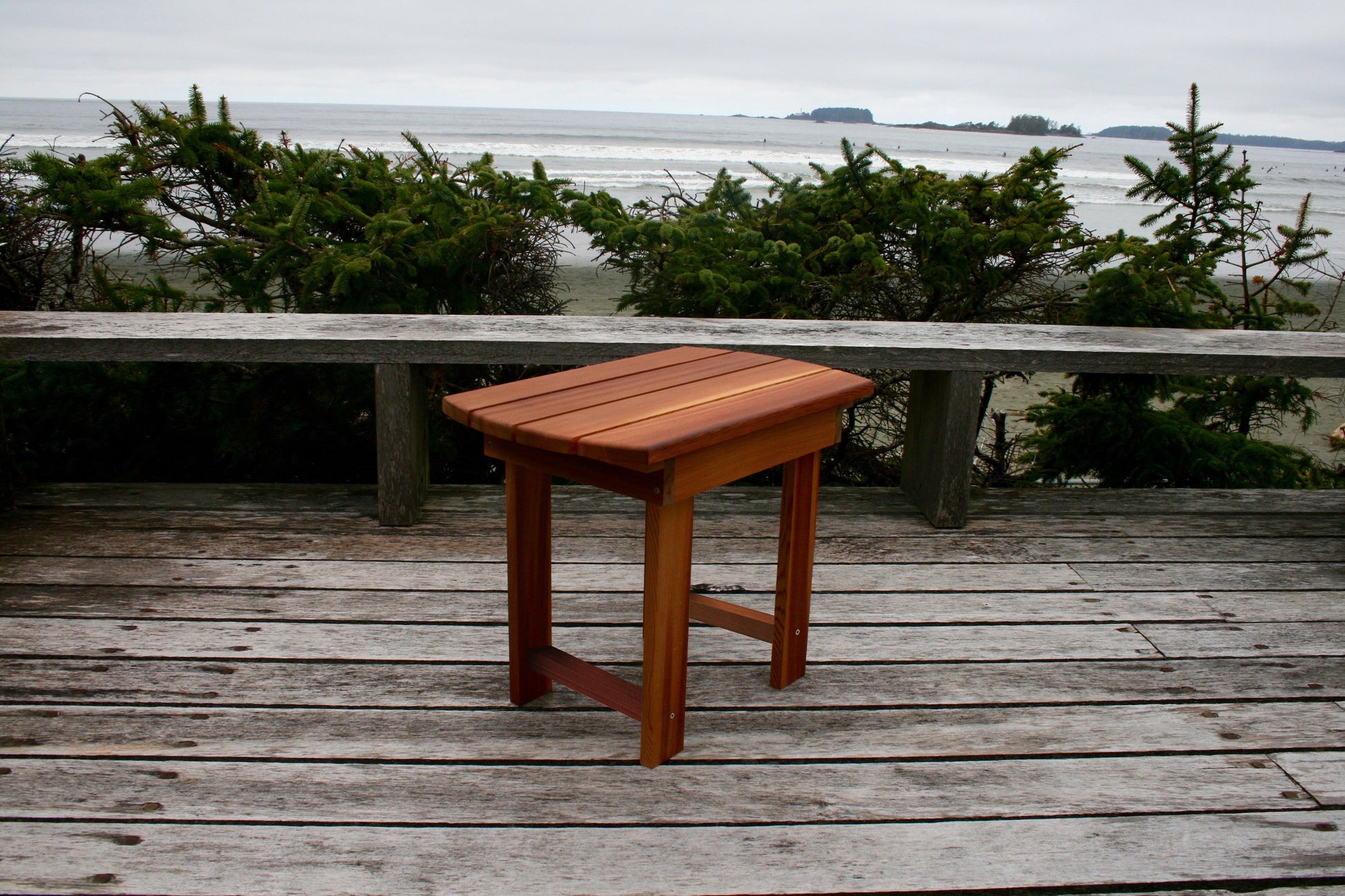 Angled view of the matching side table showing the slatted top, legs and lower support rails on a coastal deck